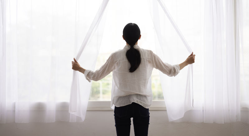 Back view of a woman opening curtains to let in morning light, representing indoor comfort and reduced outdoor noise.