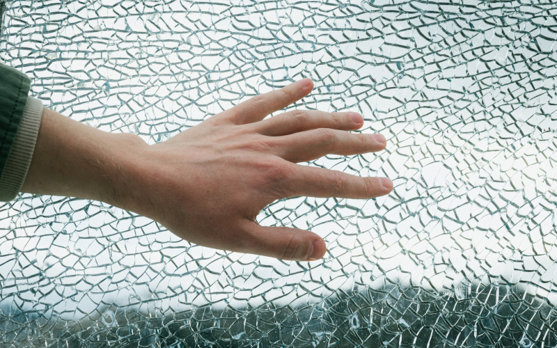 Close-up of a man’s hand touching shattered glass fragments, showing how safety film holds glass together for added protection.